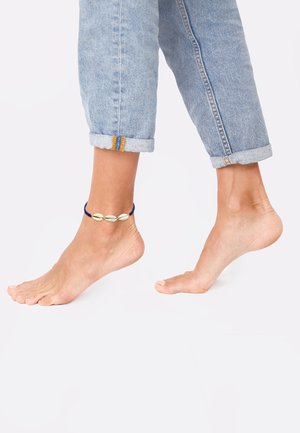 Cavigliera con cordino blu e tre conchiglie bianche, abbinata a jeans di denim azzurro arrotolati e piedi nudi.