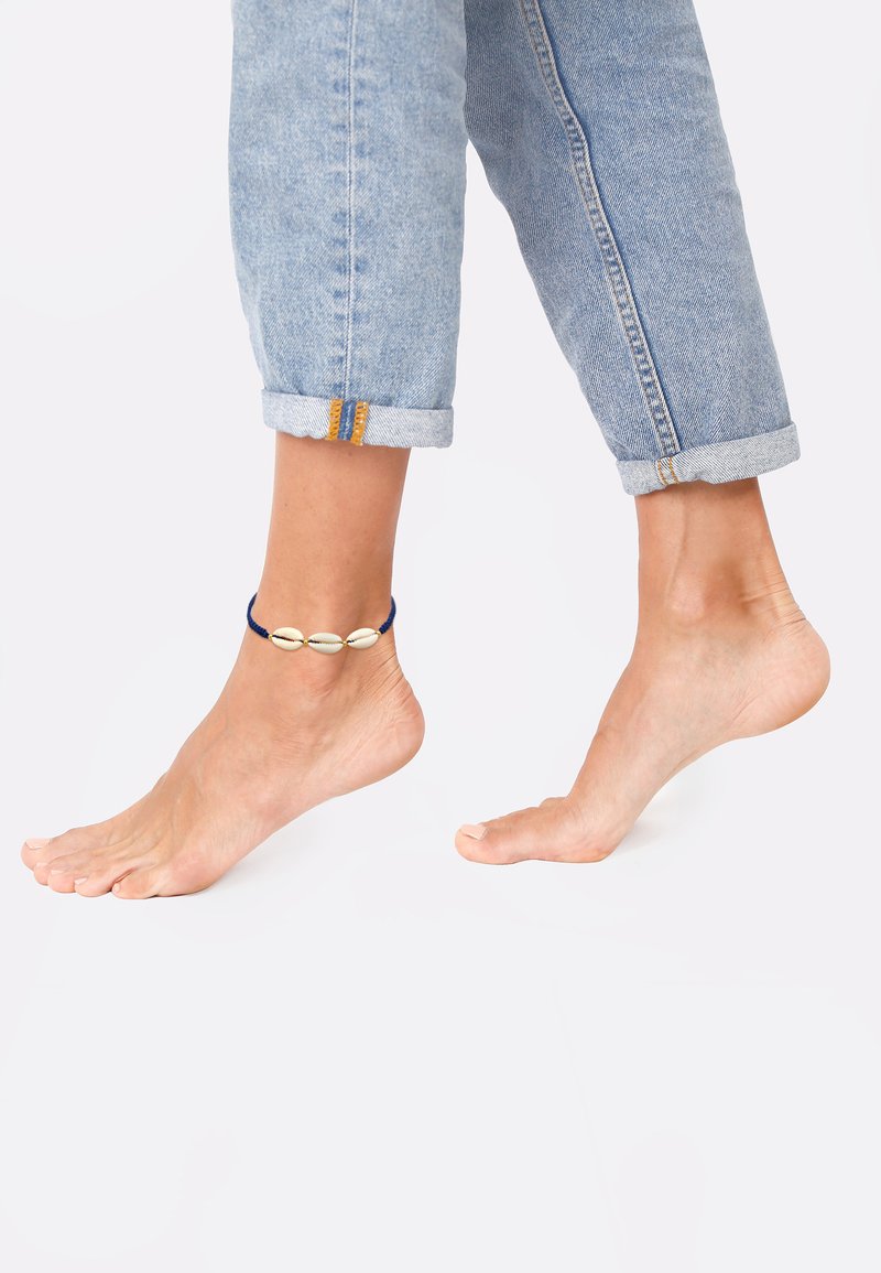 Un tobillo adornado con un cordón azul y tres conchas marinas blancas, combinado con unos pantalones de mezclilla azul claro arremangados y pies descalzos.