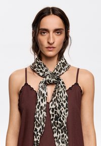 Šátek s leopardím vzorem vyrobený z lehkého materiálu, s uzlovým designem. Pozadí je bílé, model má na sobě hnědé bezrukávové tričko.