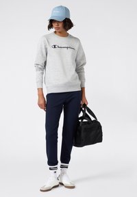 Vrouw in een grijze Champion sweatshirt, marineblauwe broek, witte sneakers, lichtblauwe pet, met een zwarte sporttas, staand tegen een witte achtergrond.