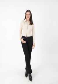 Cremfarbener, gerippter Reißverschlussoberteil mit Fronttaschen, kombiniert mit schwarzen, ausgestellten Hosen und spitzen schwarzen Stiefeln. Einfaches Design, neutrale Farben.