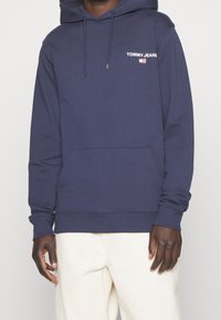 Námořnická modrá mikina s kapucí a přední kapsou, s bílým textem "TOMMY JEANS" a malým vlajkovým logem. Měkká tkanina a žebrované manžety.