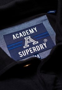Étiquette en tissu noir avec "ACADEMY SUPERDRY" en blanc et bleu, comportant un "A" stylisé et deux rayures horizontales. Indication de taille : M.
