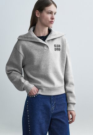 Donna che indossa una felpa grigia con la scritta "SANDRO" e collo alto, abbinata a jeans blu decorati con borchie metalliche lungo le tasche.