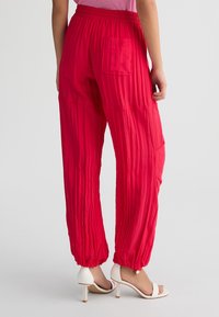 Pantaloni di tessuto rosso sgualcito con cintura elastica e tasca posteriore, affusolati alla caviglia, abbinati a sandali bianchi con tacco alto.