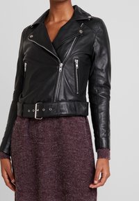 Veste de biker en cuir noir avec des poches zippées argentées, des revers larges et une taille ceinturée, portée sur un pull texturé violet.