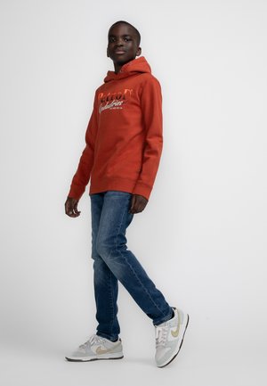 Felpa rossa con testo bianco, jeans blu e sneakers grigie Nike con dettagli dorati. La felpa ha una tasca a marsupio e cappuccio con cordoncino.