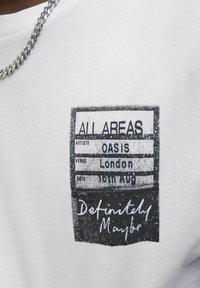 Bílý bavlněný tričko s potiskem připomínajícím koncertní vstupenku, s černým textem uvádějícím "Oasis", "Londýn" a datum.