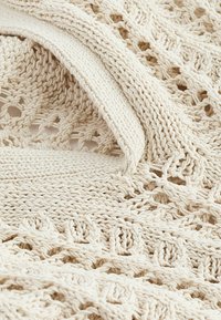 Plaid tricoté en blanc cassé avec des motifs texturés, présentant des mailles ouvertes et une bordure lisse. Matériau doux et chaud avec un design confortable.