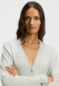 Woman with long dark hair wearing light gray cardigan, arms crossed, looking confidently at camera against plain background.