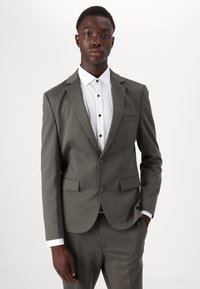 Costume gris sur mesure avec revers crantés, deux poches avant et pantalon assorti. Chemise blanche avec des boutons noirs dépassant en dessous.