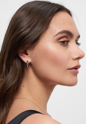 Silberne Creole mit strukturiertem Design, das überlappende Details aufweist. Das Modell hat langes Haar und trägt eine dünne Silberkette.
