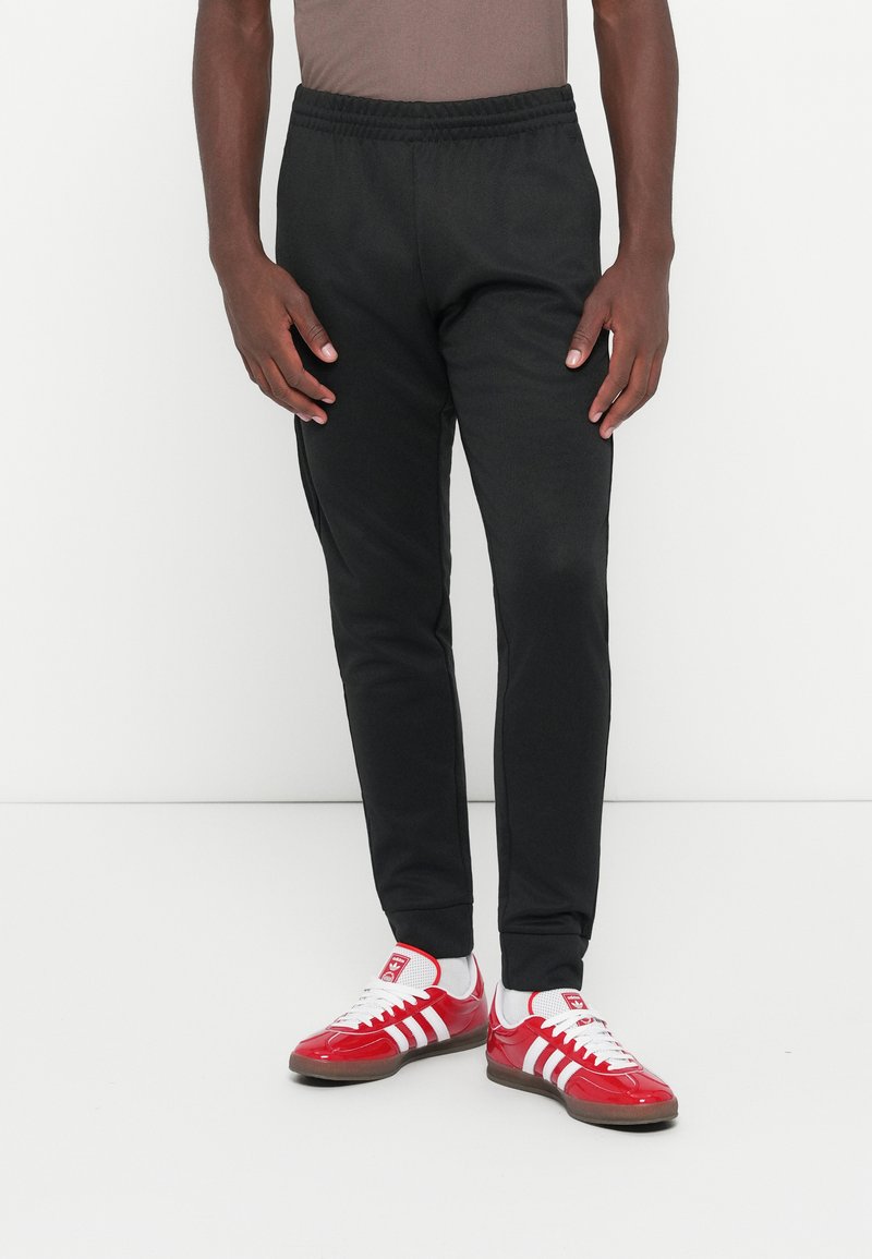 Pantaloni de trening negri cu talie elastică și șnur, cu croi ajustat și buzunare laterale, asortați cu pantofi sport roșii cu dungi albe.