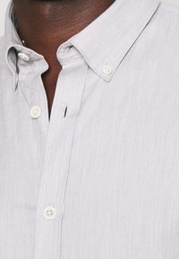 Lichtgrijze button-up shirt met een visgraatstructuur, klassieke kraag en witte knopen; zichtbaar stikseldetail nabij de knopenlijst.