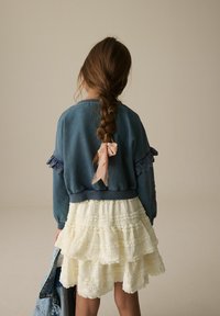Blauwe fleece sweatshirt met kantaccenten op de mouwen, gecombineerd met een meerlagige, gerimpelde witte rok. Roze lintdetail aan de achterkant.
