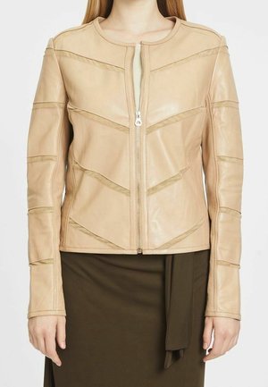 Frau trägt eine beige Lederjacke mit Reißverschluss und Chevron-Steppdetail sowie einen dunkelbraunen Rock mit gebundenem Stoffgürtel.