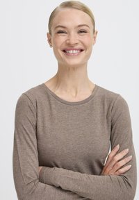 Dlhý rukáv hnedý top vyrobený z mäkkej, textúrovanej látky s okrúhlym výstrihom. Modelka má prekrížené paže a usmieva sa, na jednoduchej pozadí.