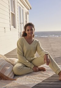 Helles gelbes, geripptes Loungewear-Set mit einem V-Ausschnitt-Oberteil und langen Ärmeln, kombiniert mit passenden Hosen. Der Hintergrund zeigt eine Holzterrasse mit Blick aufs Meer.
