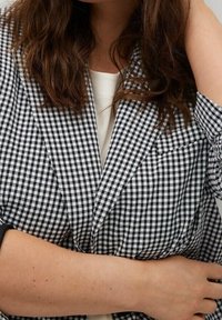 Czarny i biały blezer w kratkę z wyciętym kołnierzem, przednią kieszenią i luźnym krojem, noszony na białej bluzce, eksponujący fakturowany materiał.