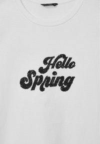 Maglietta bianca in cotone con una grafica testurizzata nera glitterata che riporta la scritta "Hello Spring" in un font stilizzato e curvo.