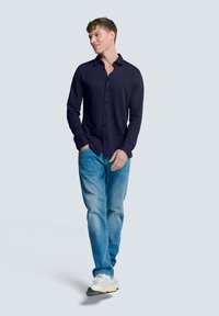 Marineblauwe overhemdd met lange mouwen van zachte stof, gecombineerd met lichtblauwe jeans en witte sneakers. Model loopt tegen een neutrale achtergrond.