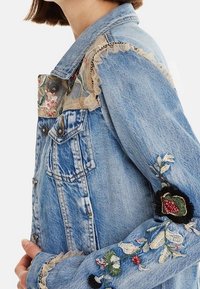 Chaqueta de denim con un lavado azul claro, adornada con bordados florales y acabados de encaje en los hombros y puños, detallada con acentos multicolores.