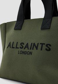 Žalia drobės pirkinių maišelio su juodomis rankenomis. Yra „ALLSAINTS LONDON“ logotipas juodu siuvinėjimu. Tekstūruota paviršius su struktūrizuotu dizainu.