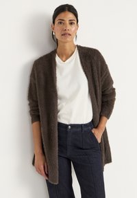 Cardigan coklat berbulu terbuat dari bahan lembut, depan terbuka, lengan panjang. Dipadukan dengan kaus v-neck putih dan celana denim gelap.