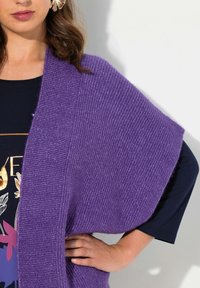 Lila stickad sjal med ribbat mönster som hänger löst över en mörk topp. Har vida ärmar och ett mjukt, mysigt utseende.