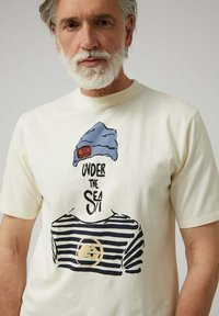 Camiseta color crema con un estampado gráfico de un gorro azul y una camiseta a rayas, con un emblema redondo en el centro.