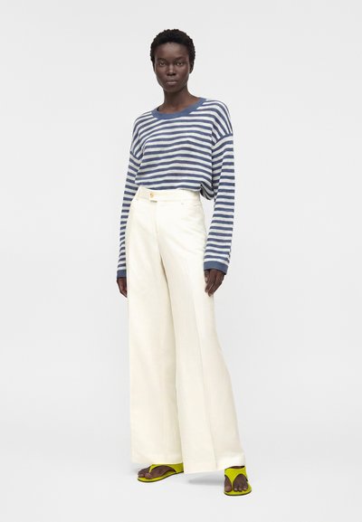 Γυναίκα που φορά μπλε και λευκή ριγέ μακρυμάνικη μπλούζα, λευκά φαρδιά παντελόνια ψηλόμεσα και κίτρινα σανδάλια με ανοιχτά δάχτυλα, στέκεται όρθια.