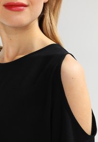 Sort kjole med skulderudskæringer, tekstureret stof, bred halsudskæring og glat, flydende design. Fremhævet med en minimalistisk stil.