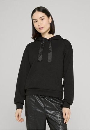 Giovane donna con capelli lisci e scuri indossa una felpa nera con cordoncini in raso e pantaloni neri in ecopelle, in piedi davanti a uno sfondo grigio chiaro.