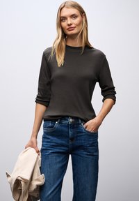 Donkergrijze lange mouwen shirt met een ronde hals, gecombineerd met mid-blauwe brede jeans. Het model houdt een lichtbeige jas vast.
