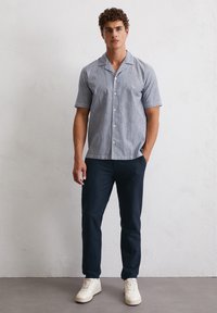 Short-sleeve, light blue-striped shirt with a camp collar, paired with dark navy pants and cream sneakers. Smooth fabric, casual fit.