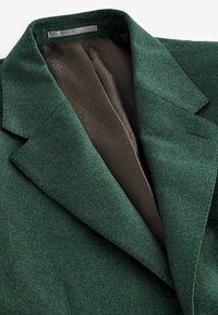 Blazer de lana verde con solapa en punta, que cuenta con un interior forrado en marrón oscuro con un patrón geométrico. Tejido texturizado y corte entallado.
