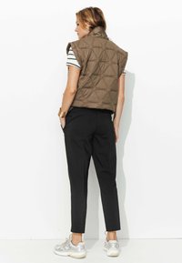 Gilet matelassé marron sans manches avec un col haut sur une chemise rayée noire et blanche, associé à un pantalon noir fuselé et des baskets argentées.
