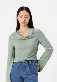 Top de manga larga con cuello drapeado en tejido de color verde claro texturizado, combinado con pantalones de mezclilla de talle alto con lazo en la parte delantera y aberturas laterales.