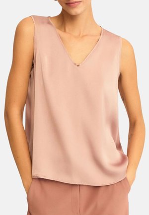 Donna che indossa una camicetta smanicata rosa chiaro con scollo a V, con le mani infilate nelle tasche dei pantaloni rosa coordinati su sfondo semplice.