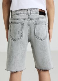Kind trägt helle Jeansshorts mit rohen Säumen und ausgebleichtem Design, von hinten vor einem schlichten Hintergrund gezeigt.