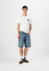Hvit t-skjorte med kort erme og svart rundt logo, kombinert med knelange lyseblå denimshorts og svarte joggesko med hvite snøringer.