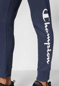 Marina blå mjukisbyxor gjorda av en bomullsblandning med ribbade muddar, med en vit "Champion"-logotyp tryckt vertikalt längs sidan.