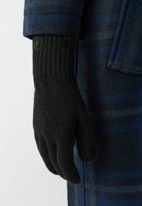 Juodos megztos pirštinės su banguota apykakle ir apvaliu akcentiniu mygtuku, dėvimos prieš mėlynai-pilkos raštuotos striukės.