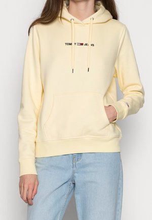 Personne portant un sweat à capuche Tommy Jeans jaune pâle avec une poche avant et un jean bleu clair sur un fond uni.