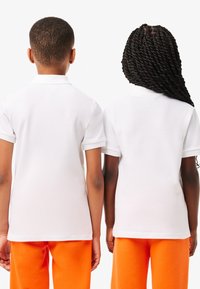 Twee kinderen staan met hun rug naar de camera, gekleed in witte poloshirts en feloranje broeken, tegen een egale achtergrond.