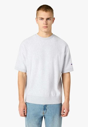 Giovane uomo con capelli corti che indossa una felpa grigio chiaro a maniche corte e jeans blu, in piedi davanti a uno sfondo bianco uniforme.