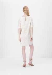 Abito bianco in pizzo con motivi floreali, maniche corte e una apertura circolare sul retro. Abbinato a sandali con tacco a strappo marrone chiaro.