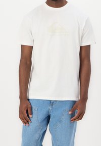 Man med en enkel vit Quiksilver t-shirt och ljusblå jeans, som står mot en vit bakgrund.