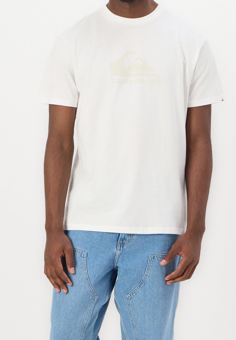 Man med en enkel vit Quiksilver t-shirt och ljusblå jeans, som står mot en vit bakgrund.