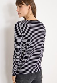 Kobieta widziana z tyłu, ubrana w granatową i białą w paski bluzkę z długim rękawem oraz ciemnoszare dżinsy, patrząca w lewo na tle jednolitego białego tła.